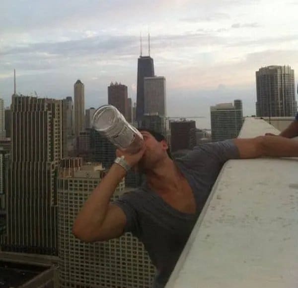 Man chugging from a large bottle while leaning over a high rooftop ledge above Chicago.