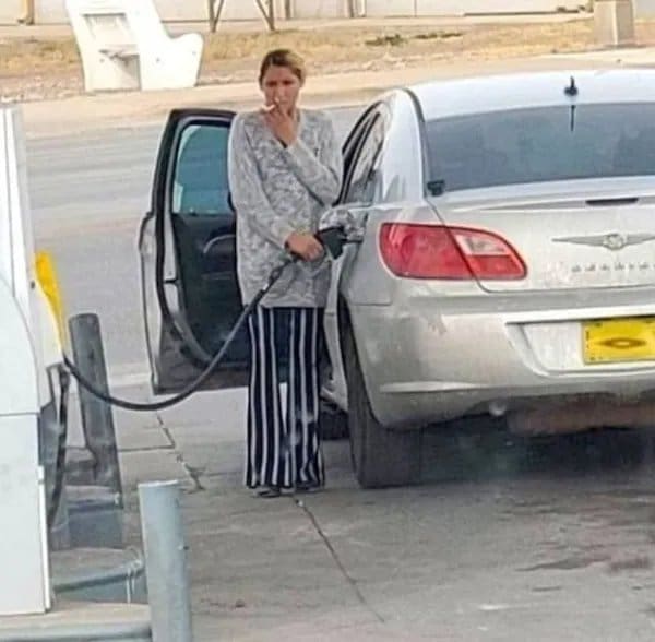Woman smoking a cigarette while pumping gas into a silver sedan at a gas station.