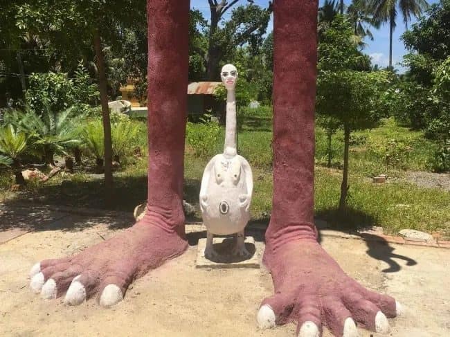 Strange white long-necked statue standing between two giant pink concrete feet in a garden.