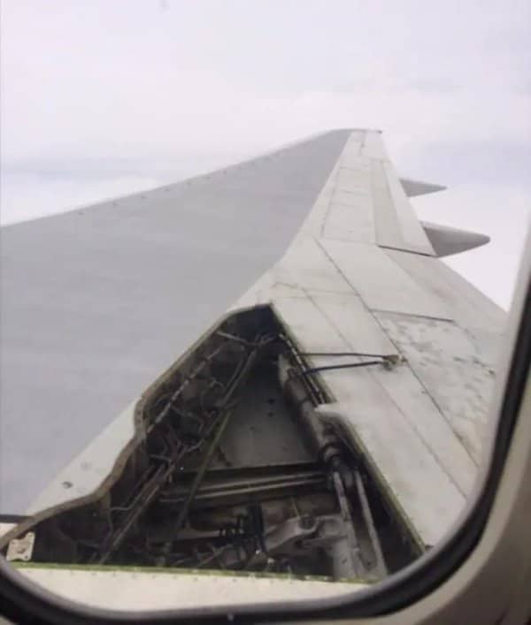Airplane window view of a wing with a missing panel exposing wires and machinery