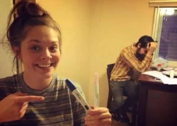 Smiling woman holding a positive pregnancy test while a man behind her clutches his head