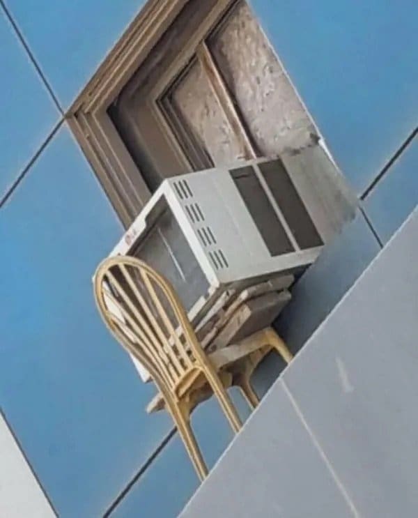 Window air conditioner balanced on a wicker chair outside a high apartment window.
