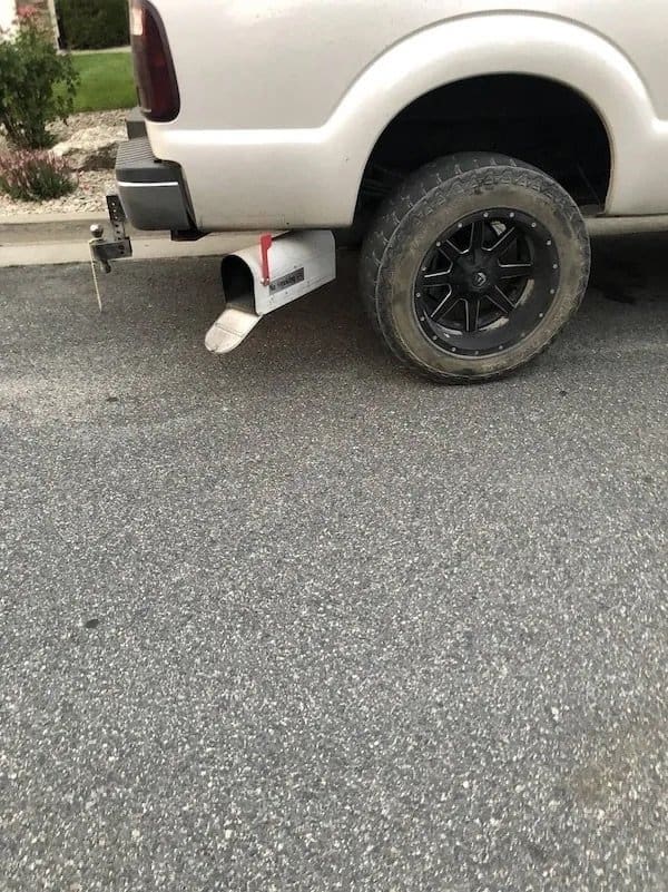 Metal mailbox mounted as an exhaust tip under the back of a white pickup truck.