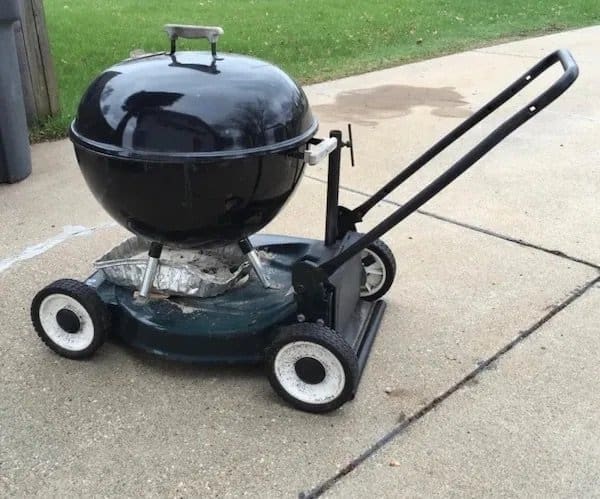 Black kettle grill mounted onto an old push lawn mower base with wheels.