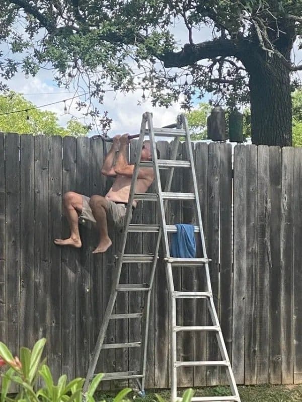 Shirtless man doing pull-ups on a bar between a ladder and a backyard fence.