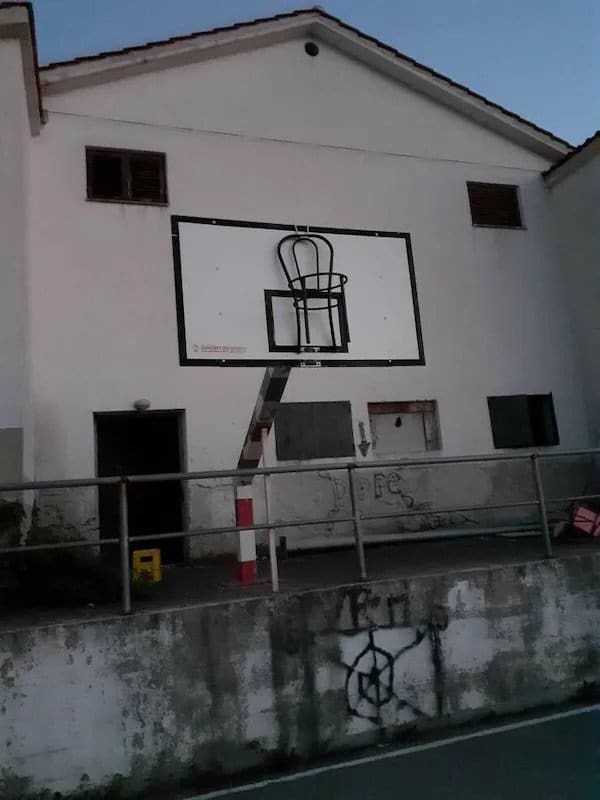 Outdoor basketball backboard with a wooden chair hung up in place of the rim.