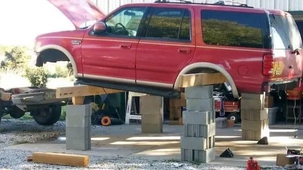 Red SUV propped up on stacked cinder blocks and lumber for a driveway repair.