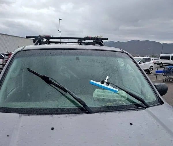 Blue window squeegee zip tied to a car windshield wiper blade in a parking lot.