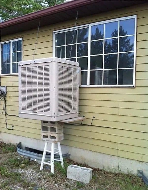 Swamp cooler balanced on cinder blocks and a small white stool beside a house window.