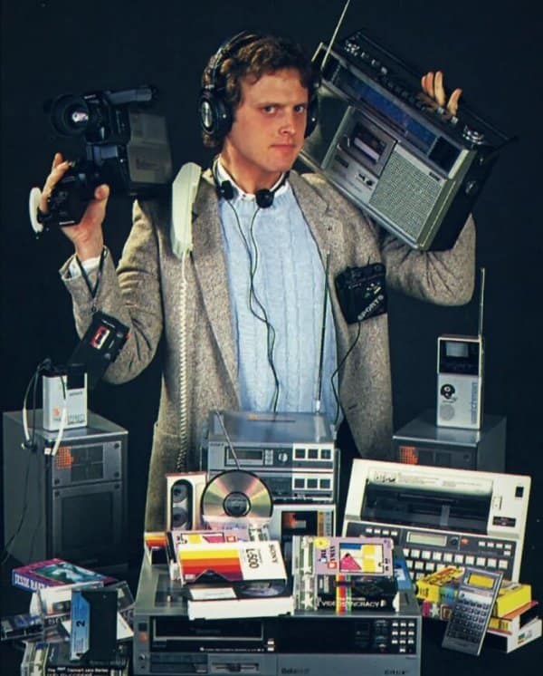 1980s man overburdened with dozens of electronic devices including boomboxes, camcorders, Walkmans, and VCRs illustrating retro tech.