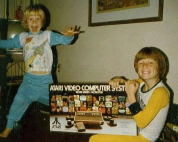 Vintage home photo of two ecstatic kids in pajamas posing with a boxed Atari Video Computer System in a retro tech memory.