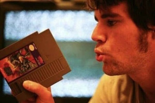 Guy blowing directly into a gray NES game cartridge to clear dust in front of a fuzzy static television screen in a retro tech ritual.