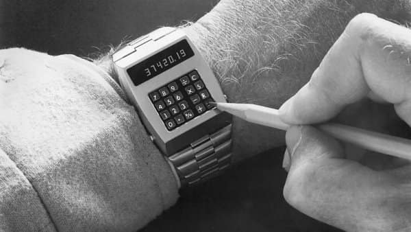 Vintage black-and-white photograph of a hand using a pencil tip to operate tiny buttons on an LED calculator wristwatch as retro tech.