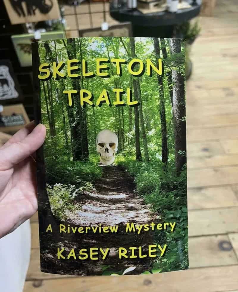 Mystery novel Skeleton Trail with a clip art skull floating over a sunny forest path.