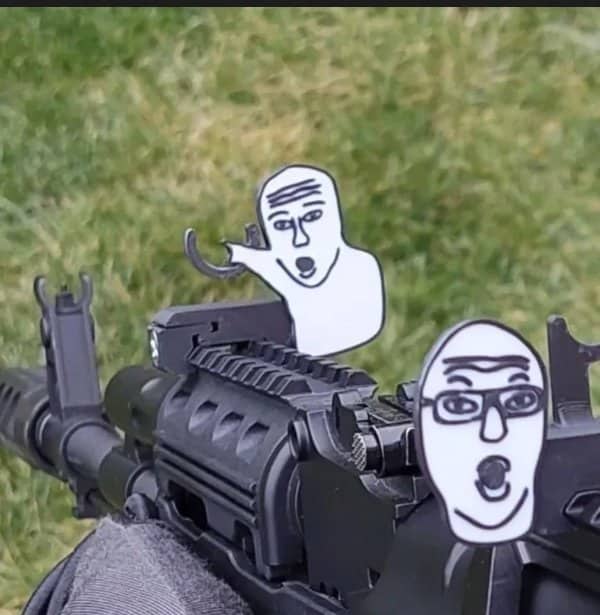 Rifle fitted with cartoon meme face cutouts on its sights, held over a grassy field