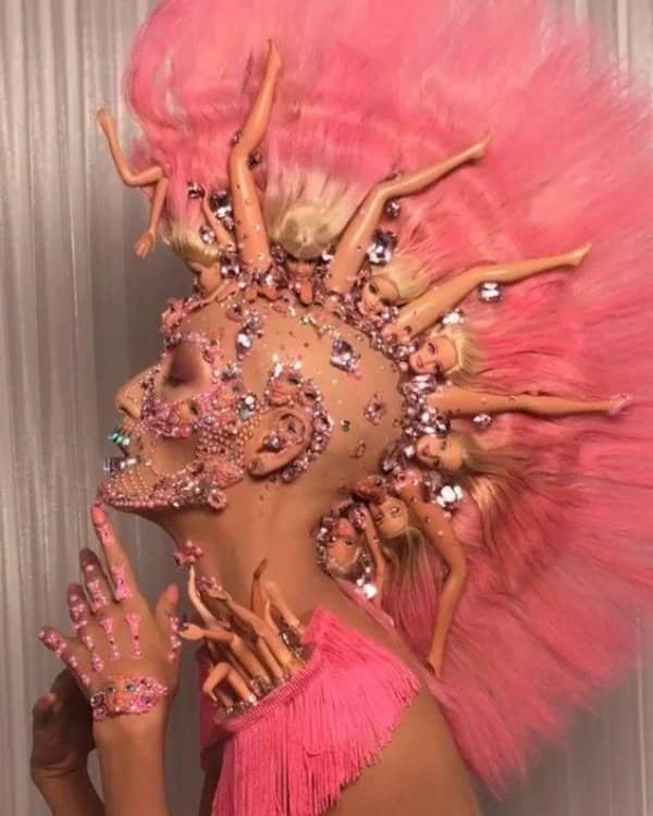 Model wearing a giant pink wig with Barbie dolls woven through it and rhinestone makeup