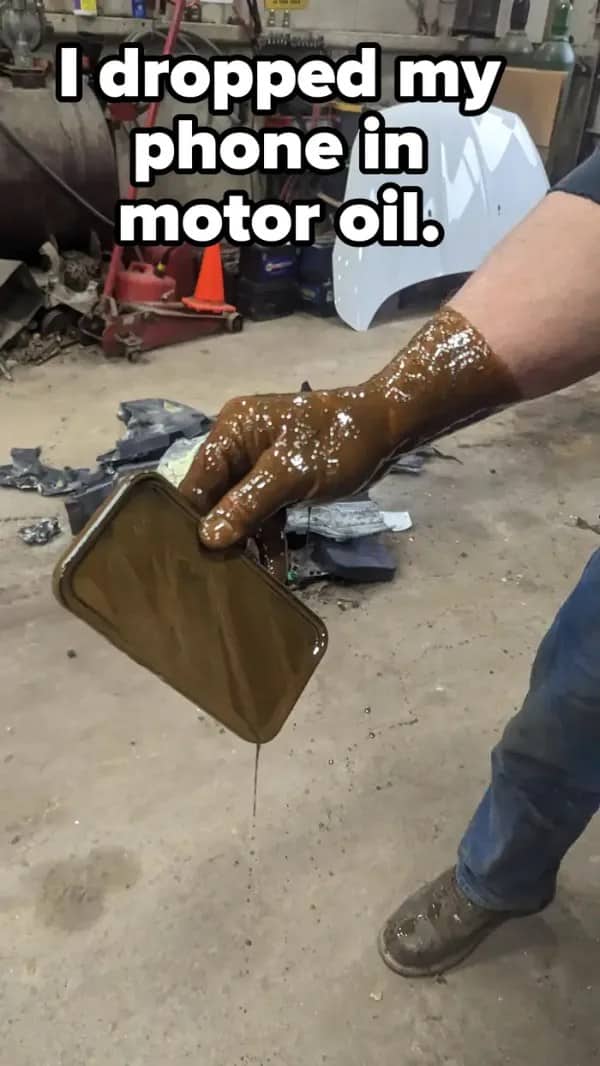Gloved hand lifting a phone coated in dripping motor oil inside a garage