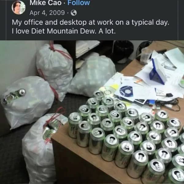 Office desk covered in rows of empty Diet Mountain Dew cans beside bags of more cans