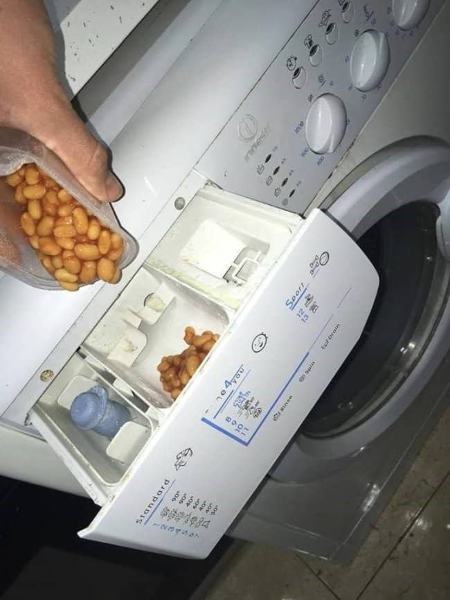 Hand pouring baked beans into the detergent drawer of a white washing machine.