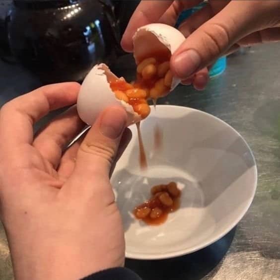 Hands cracking open an eggshell to reveal baked beans pouring into a white bowl.