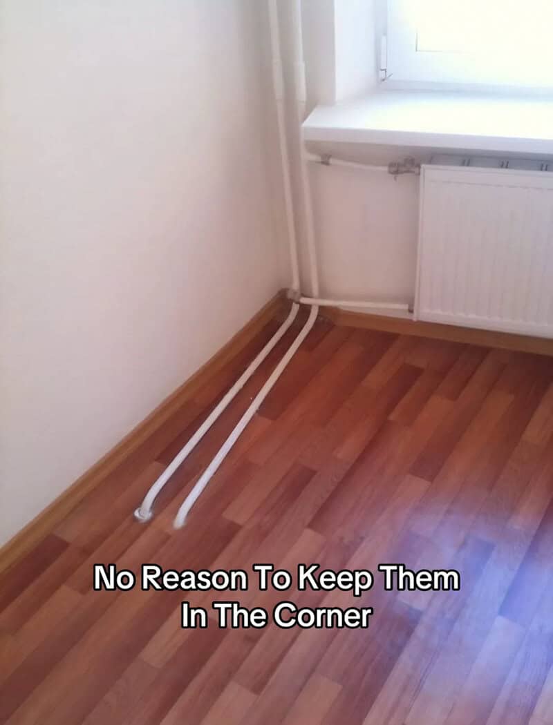 Two white heating pipes running diagonally across a wood floor away from the wall corner.