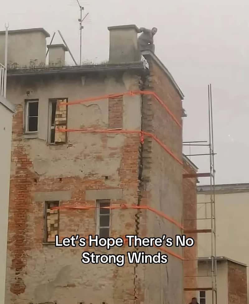 Badly cracked brick building wrapped in orange ratchet straps while a worker sits on the roof.