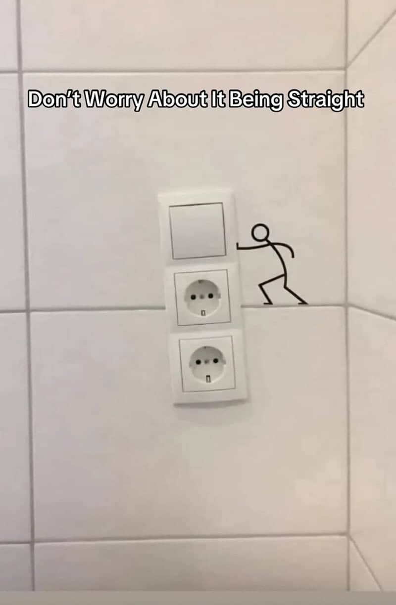 Stacked light switch and outlets installed crooked on a tile wall, with a doodled stick figure pushing.