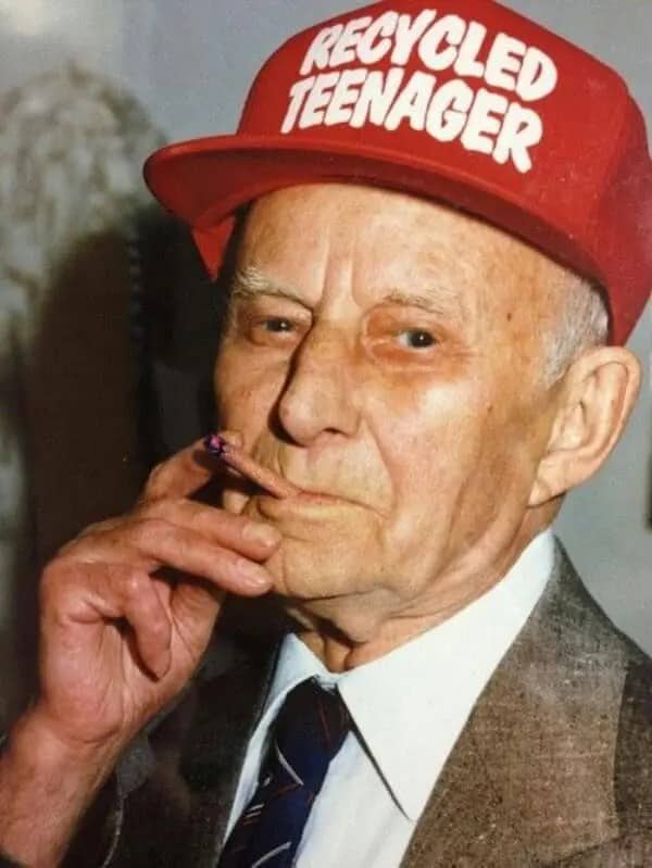 Elderly man in a suit and tie wearing a red Recycled Teenager cap while smoking.