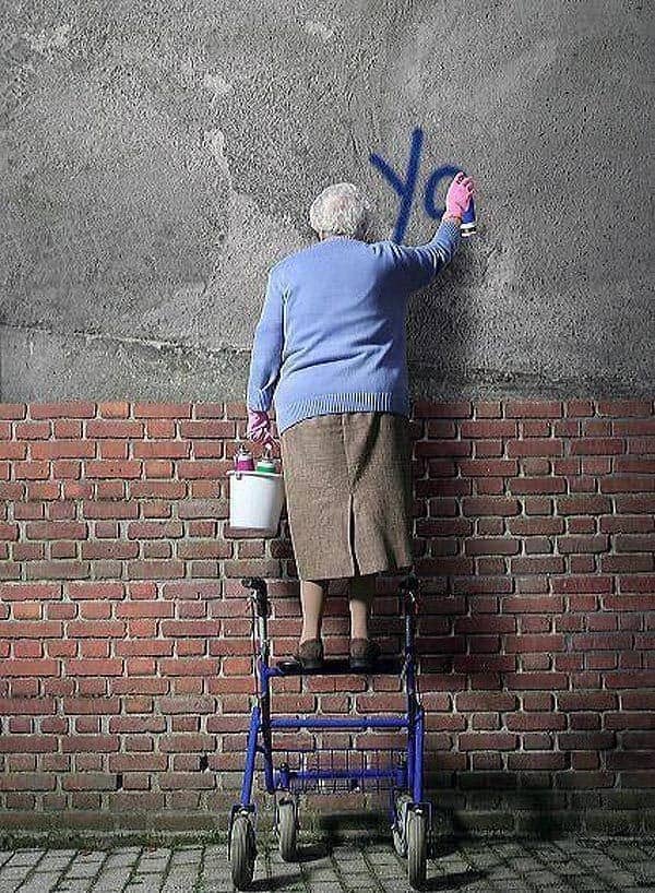 Elderly woman standing on a rolling walker to spray paint graffiti on a city wall.
