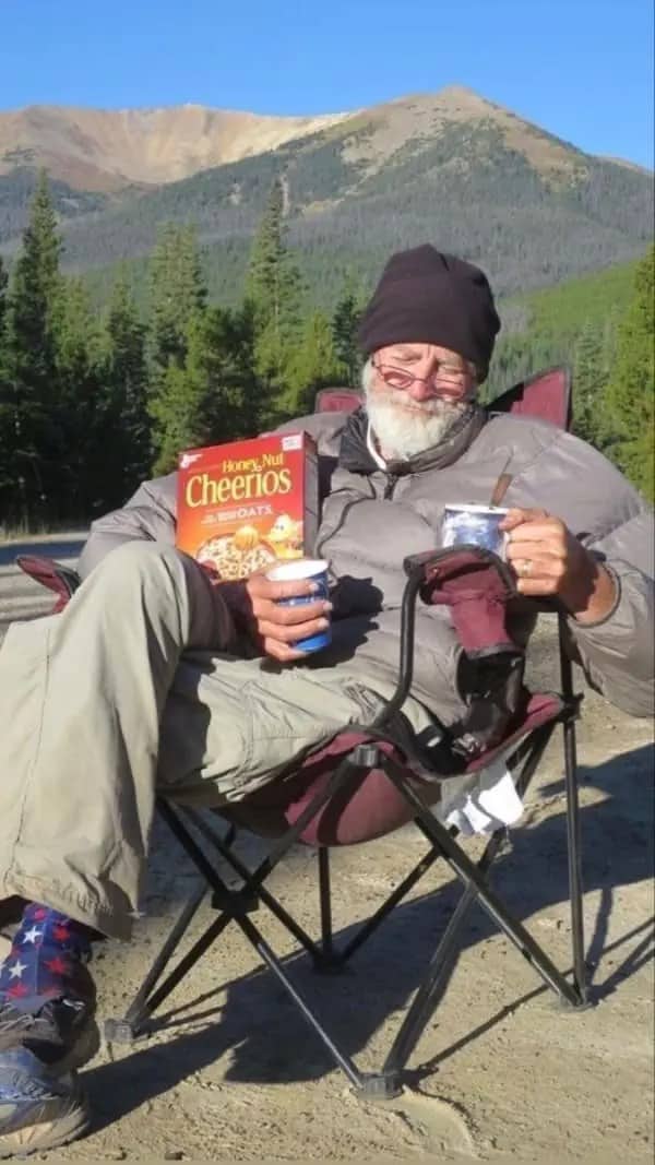 Bearded older man relaxing in a camp chair with Cheerios and coffee in front of mountains.