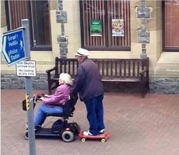 Older man on a skateboard holding onto his wife's mobility scooter on a town sidewalk.