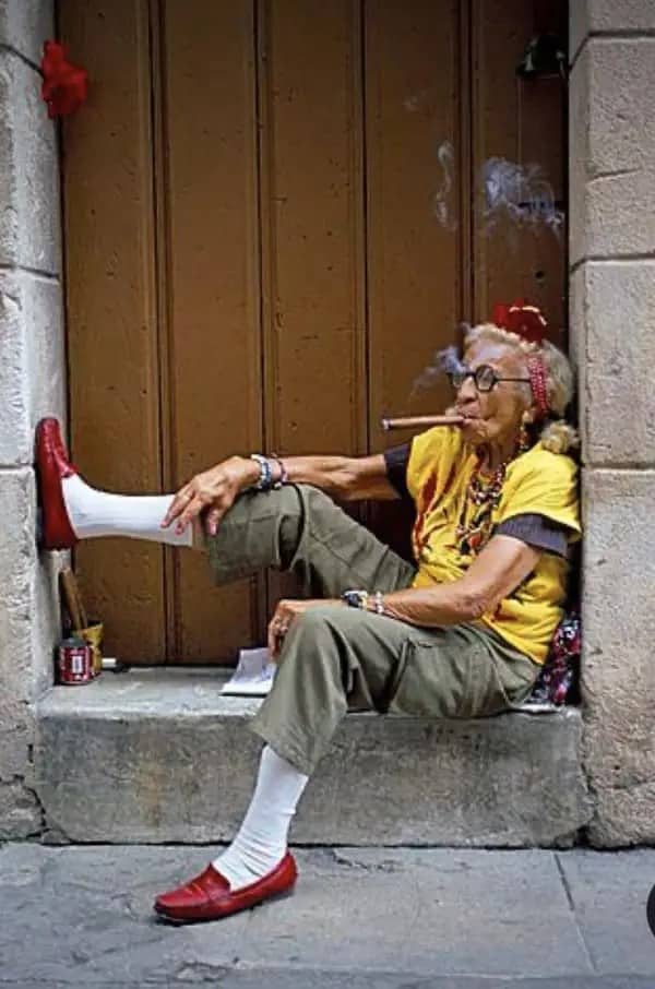 Elderly woman in red shoes and a yellow top smoking a cigar on a stone doorstep.