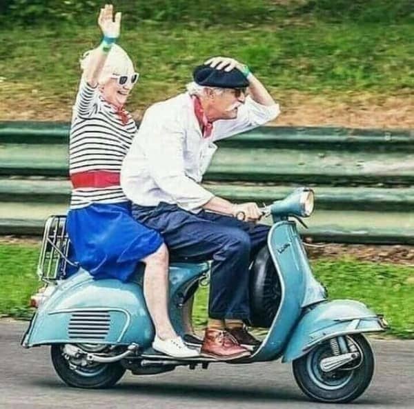 Older couple riding a vintage blue Vespa, the woman waving joyfully from the back seat.