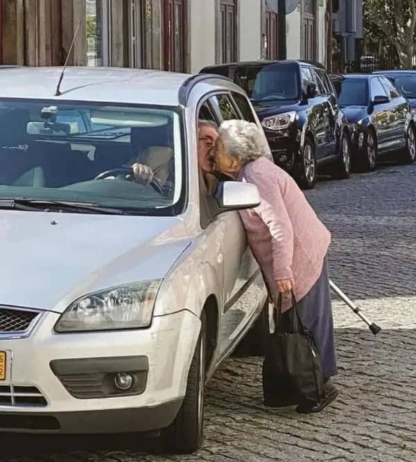 Elderly woman with a cane leaning into a car window to kiss the driver goodbye.