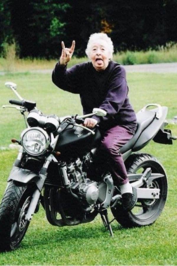 Elderly woman sitting on a motorcycle, sticking out her tongue and throwing a rock hand sign.
