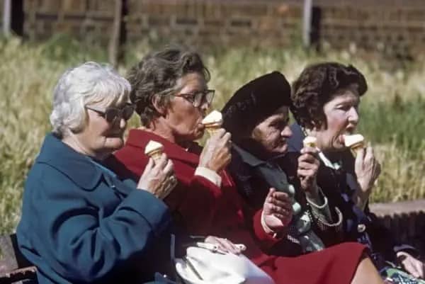 Four older women in coats eating ice cream cones together on a sunny bench.