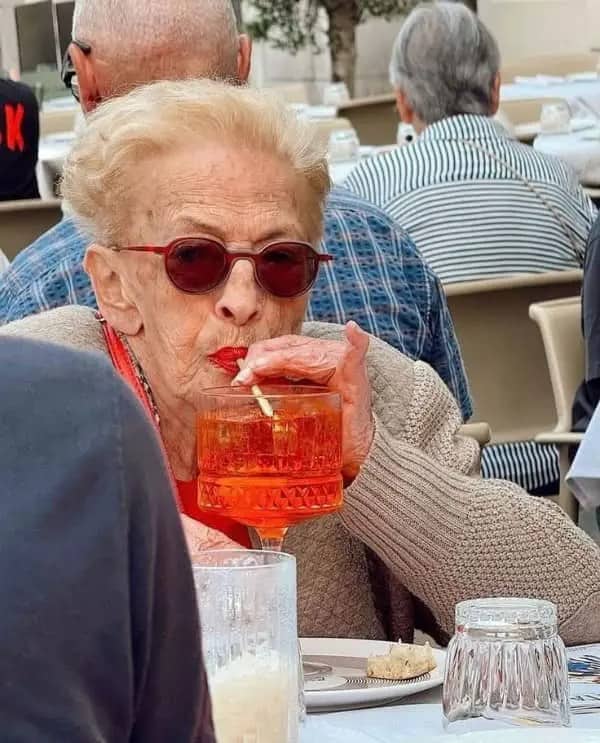 Elderly woman in red sunglasses and lipstick sipping a spritz cocktail at an outdoor cafe.