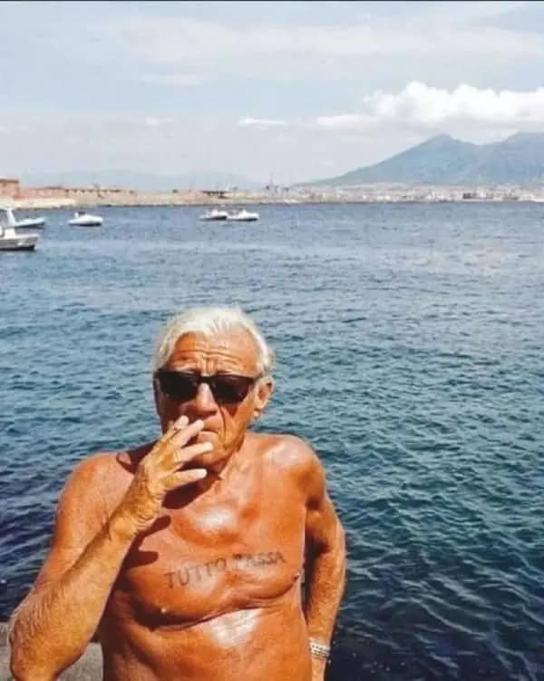 Tanned older man smoking by the sea with Tutto Passa tattooed across his chest.