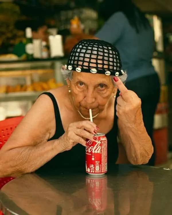 Elderly woman in a crochet cap sipping Coca-Cola through a straw while making a rude gesture.