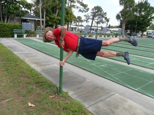Smiling older man in a red shirt holding himself sideways off a pole by a shuffleboard court.