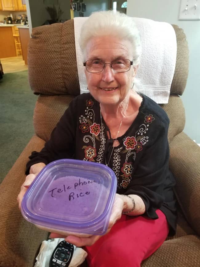 Smiling elderly woman in a recliner holding a plastic container labeled telephone rice.