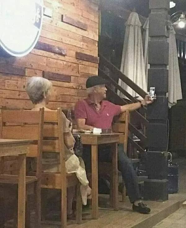 Older man in a flat cap taking a selfie at a bar table while his companion looks away.