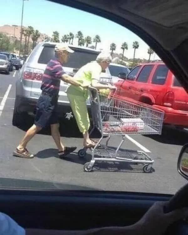Older man pushing a shopping cart while his wife rides on the back across a parking lot.