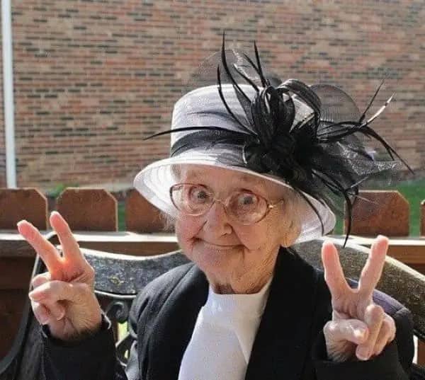 Elderly woman in a feathered black and white hat flashing double peace signs with a grin.