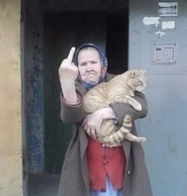 Elderly woman in a headscarf holding a ginger cat while making a rude gesture at the camera.