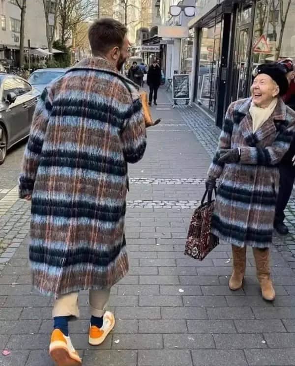 Young man and elderly woman laughing as they pass on the street in nearly identical coats.