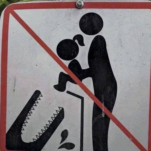 Warning sign showing a crossed-out adult dangling a child over a crocodile's open jaws.
