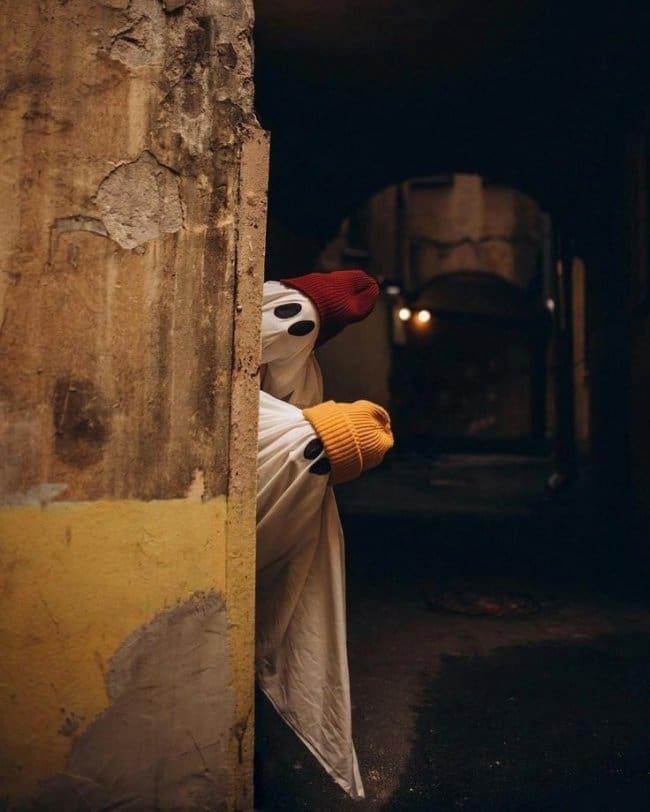 Two people in bedsheet ghost costumes with beanies peeking out from behind a wall.