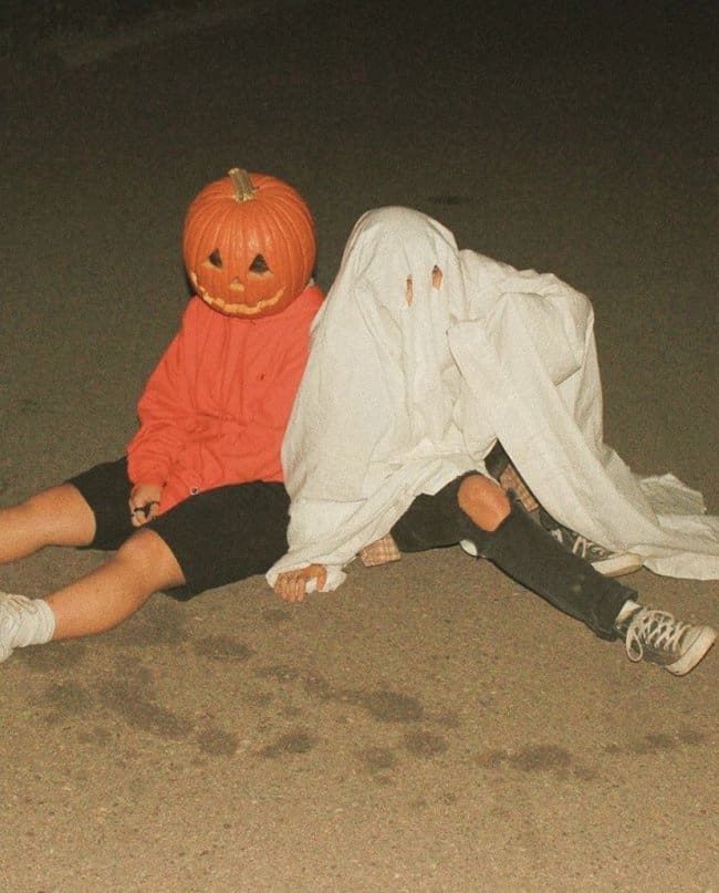 Person wearing a carved pumpkin head sitting with a friend dressed as a sheet ghost.