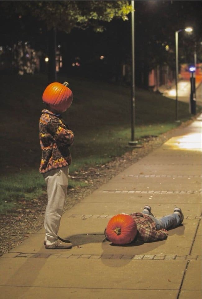Person with a pumpkin head standing over another pumpkin-headed person lying on a sidewalk.